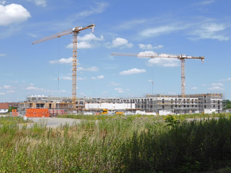 Neubaugebiet im Bau mit zwei Turmdrehkränen und eingerüsteten Rohbauten hinter einer Brachwiese