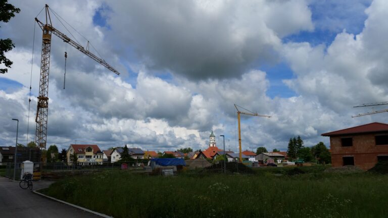 Neubaugebiet mit Rohbau eines Hauses, zwei Baukränen und Wohnhäusern unter Wolkenhimmel